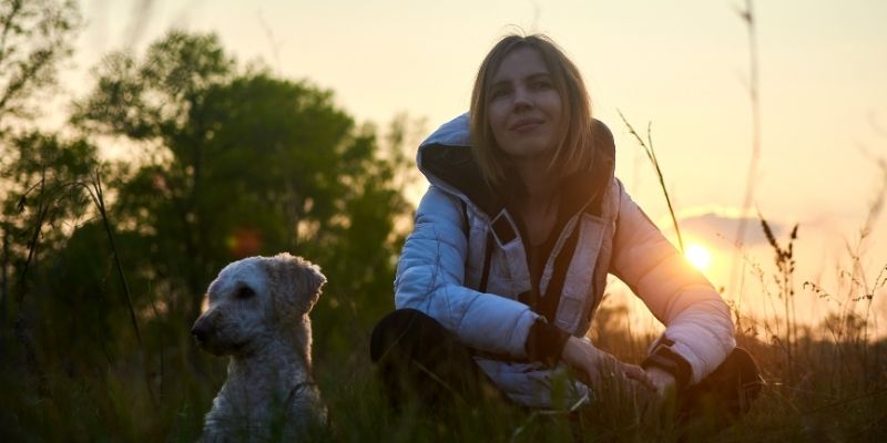 A woman sitting with her dog in quiet nature illuminated by sunbeams, symbolizing healing and the family systemic connection