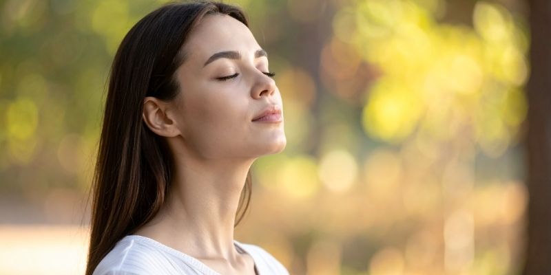 A young woman with eyes closed breathing deeply in nature to calm her thoughts and emotions