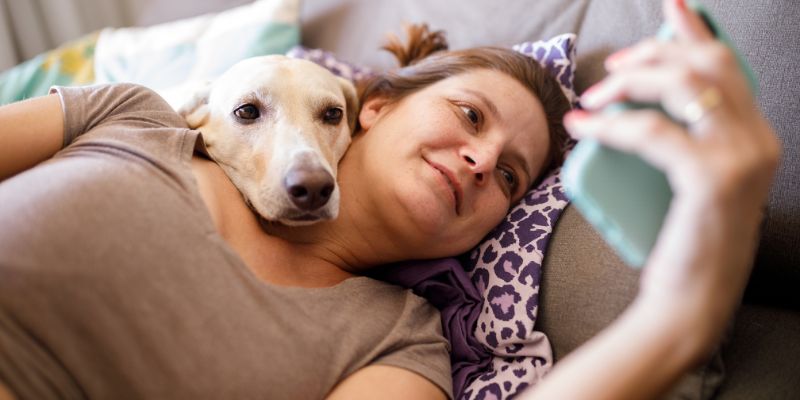 A young woman lying on a couch with her tan dog while using a smartphone to access educational resources for choosing and raising a dog