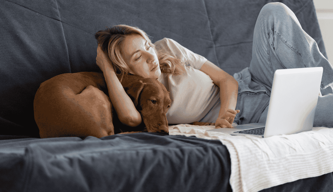 A woman lying on a dark couch with her laptop while hugging her brown dog representing the dog as a mirror of hidden emotions
