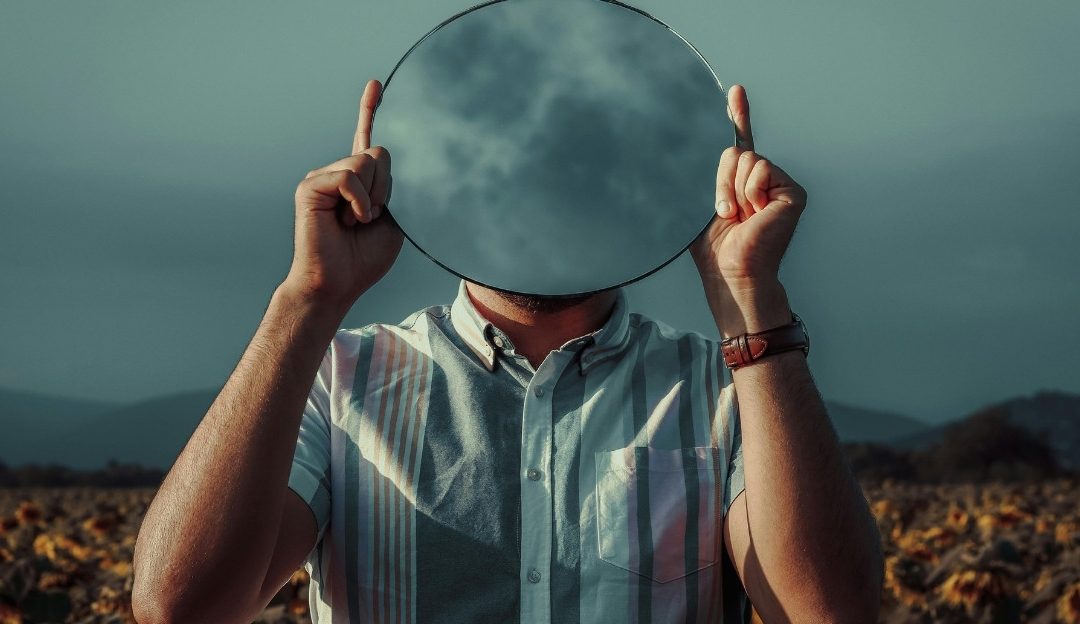 A man holding a round mirror in front of his face reflecting the sky and questioning his identity